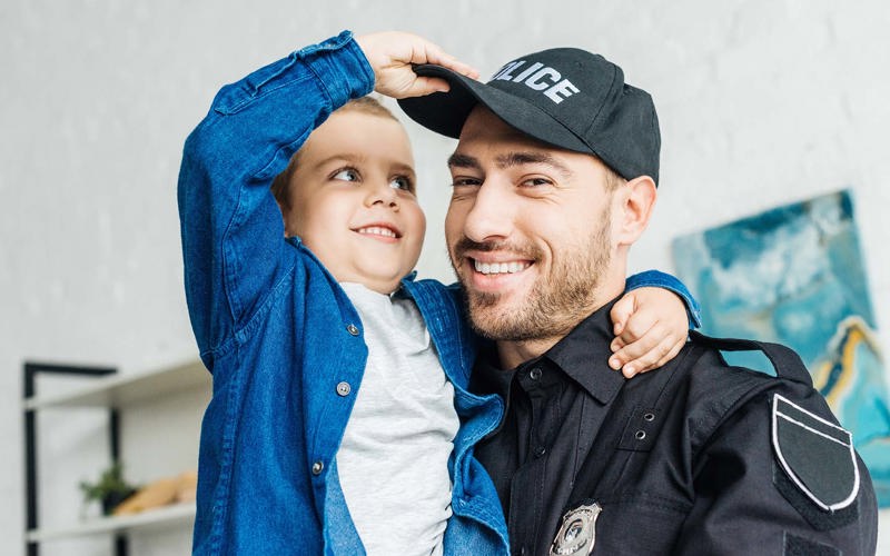 close-up portrait of smiling young father in police uniform carrying his little son and looking at camera