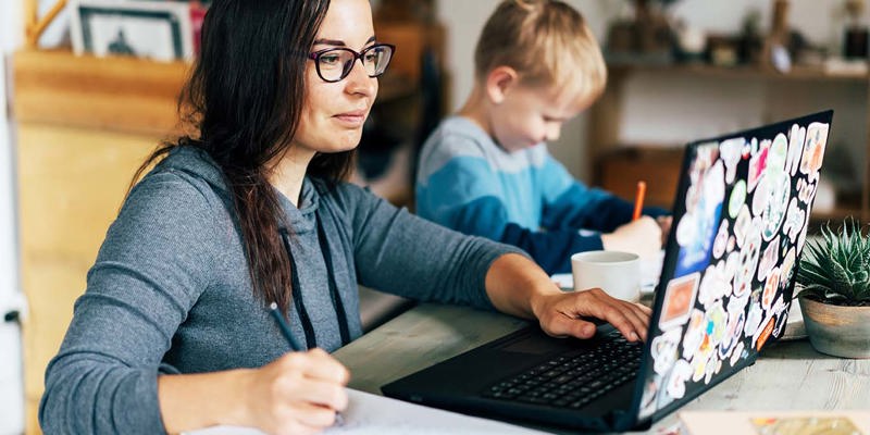 Concept of work from home and home family education. Mom and son are sitting at the desk. Business woman works on the Internet in a laptop, a child writes in a notebook.