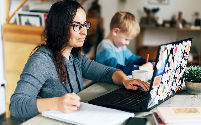 Concept of work from home and home family education. Mom and son are sitting at the desk. Business woman works on the Internet in a laptop, a child writes in a notebook.