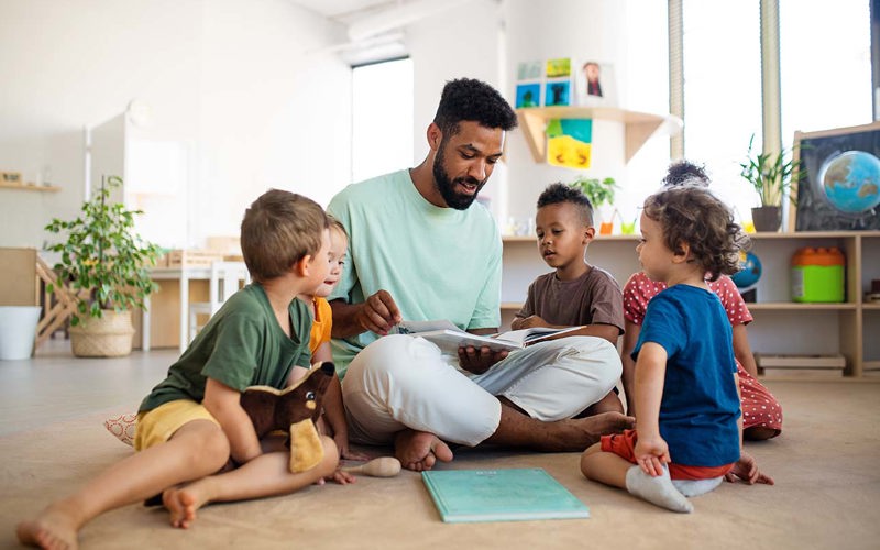 Group of small nursery school children with man teacher sitting on floor indoors in classroom, having lesson.