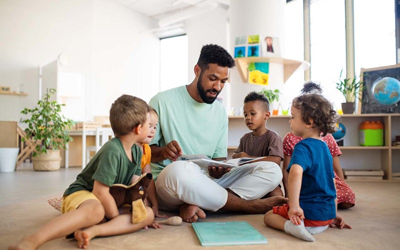 Group of small nursery school children with man teacher sitting on floor indoors in classroom, having lesson.