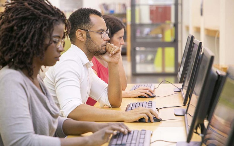 Multiracial group of students training in computer class. Line of man and women in casual sitting at table, using desktops, typing, looking at monitor. Training center concept.