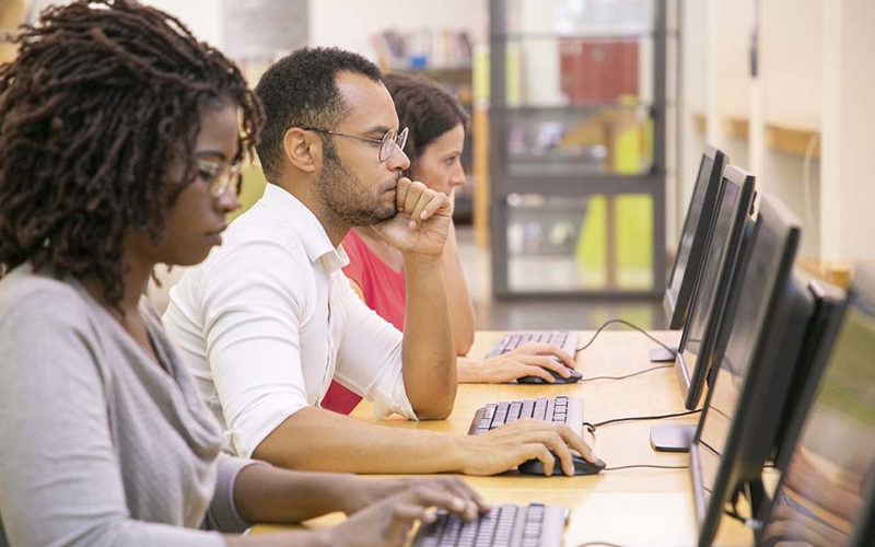 Multiracial group of students training in computer class. Line of man and women in casual sitting at table, using desktops, typing, looking at monitor. Training center concept