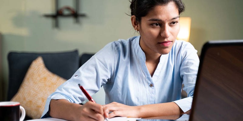 Young business woman writing down notes by looking laptop - concept of employee or student online training class at home.